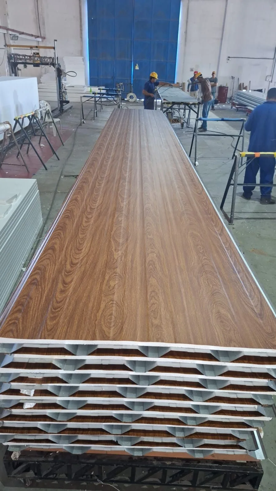 Equipe da Ecorenovar produzindo painel de telha termoacústica com acabamento amadeirado na linha de fabricação, painéis empilhados com núcleo isolante visível
