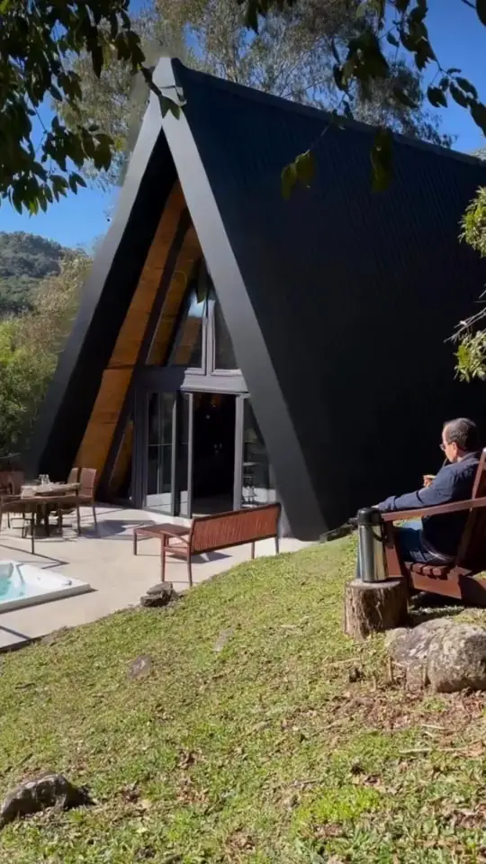 Chalé A-frame com cobertura metálica escura e fachada de vidro, em meio ao verde